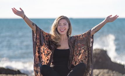 Person sitting on sand with arms raised, smiling, near the ocean with rocks in the background, clear sky.