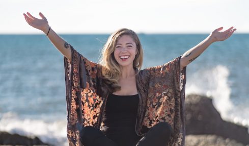 Person sitting on sand with arms raised, smiling, near the ocean with rocks in the background, clear sky.