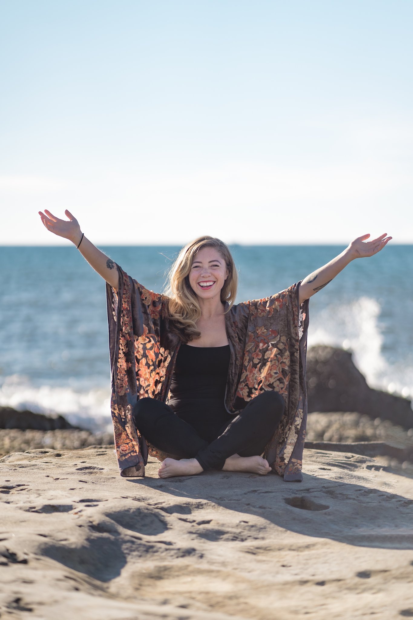 Person sitting on sand with arms raised, smiling, near the ocean with rocks in the background, clear sky.