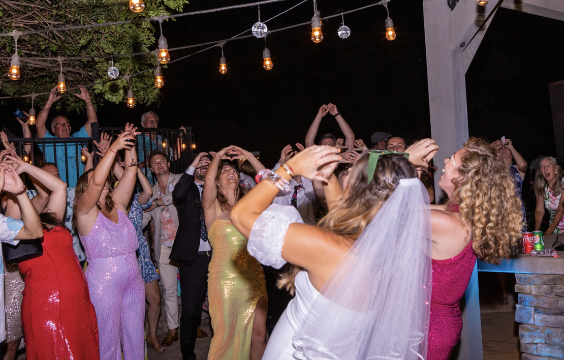 People dancing at a party under string lights, some with arms raised, wearing colorful dresses and a bride with a veil.