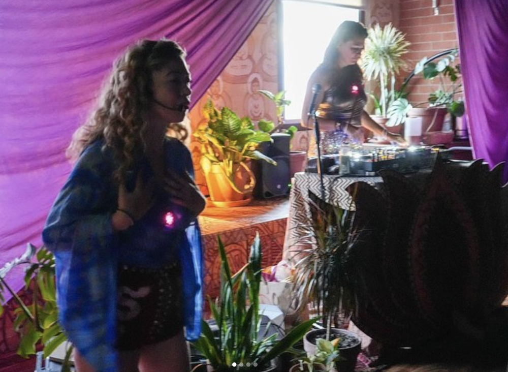 Two women in a room with purple curtains, plants, and a DJ setup, one in the foreground and one at the DJ table.