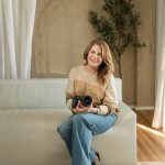 Woman sitting on a sofa holding a camera, smiling, in a room with a tree and curtains.
