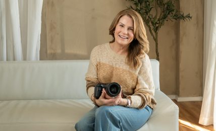 Woman sitting on a sofa holding a camera, smiling, in a room with a tree and curtains.