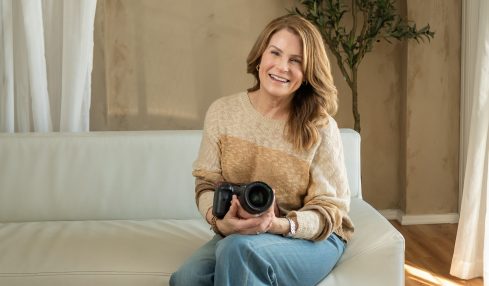 Woman sitting on a sofa holding a camera, smiling, in a room with a tree and curtains.