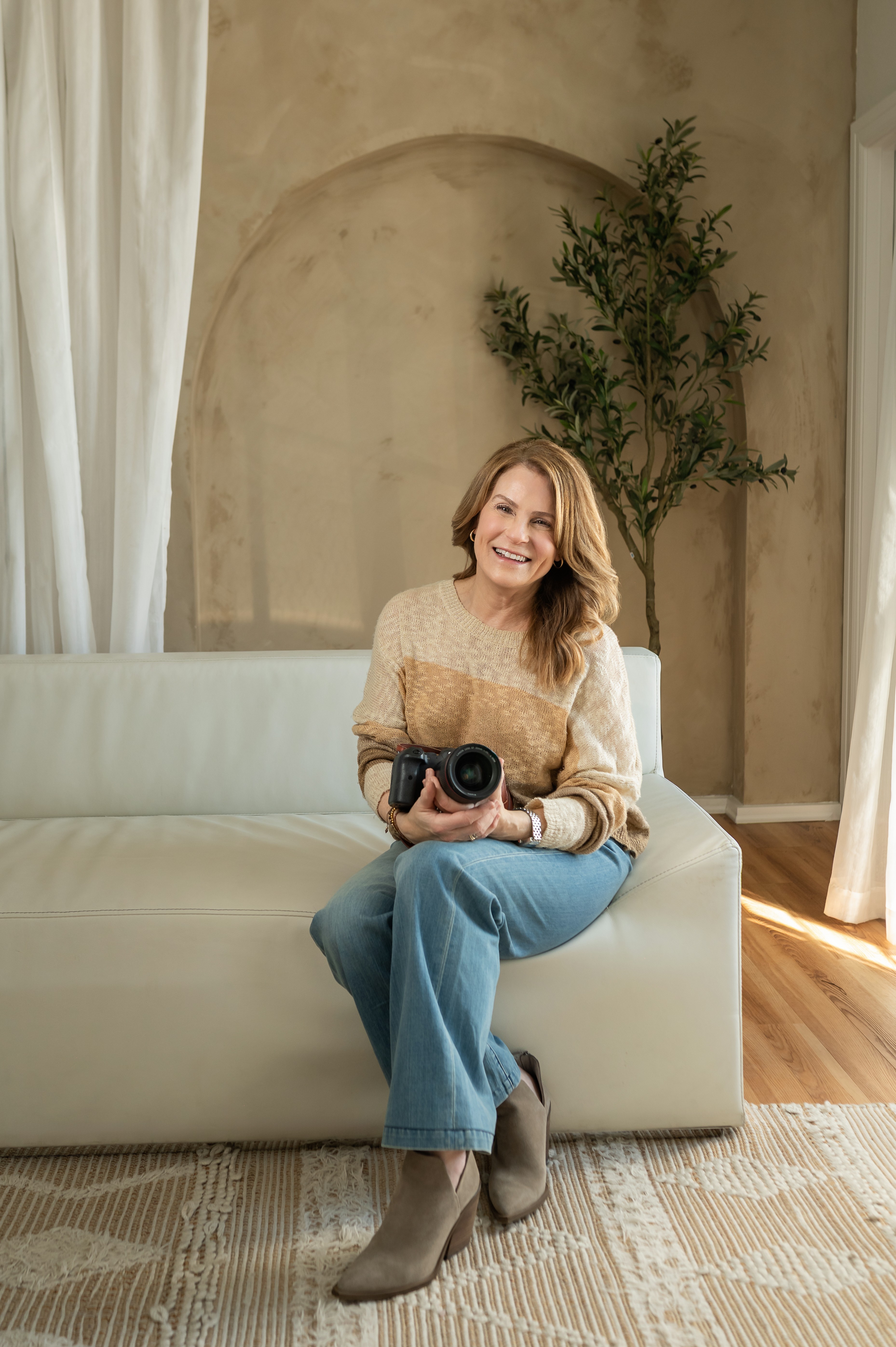 Woman sitting on a sofa holding a camera, smiling, in a room with a tree and curtains.