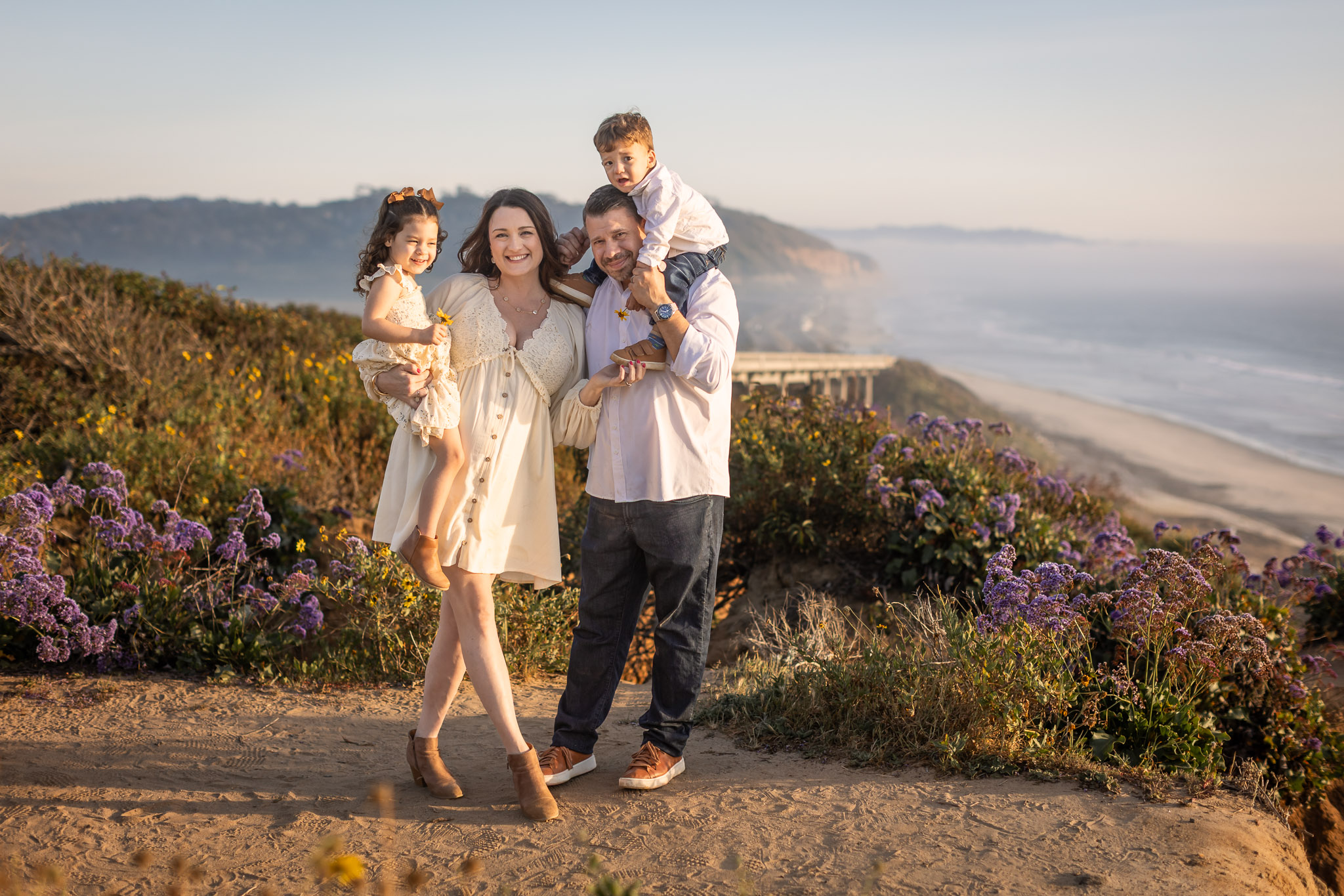 Family of four standing outdoors near a beach with purple flowers, smiling, with ocean and cliffs in background.