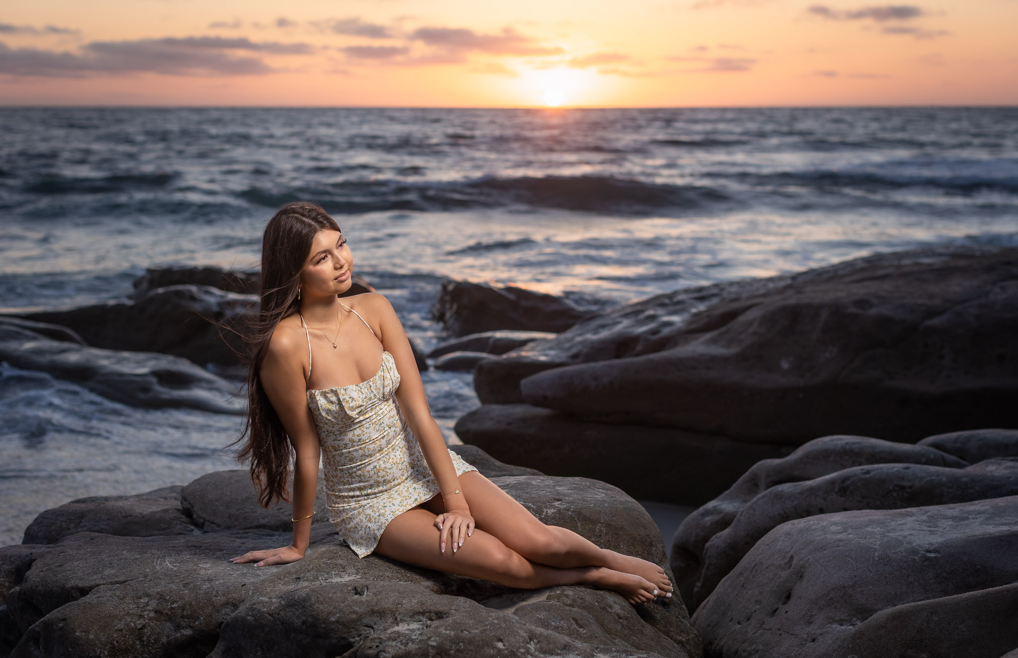 Woman with long hair sitting on rocks by the ocean at sunset, wearing a light-colored dress.