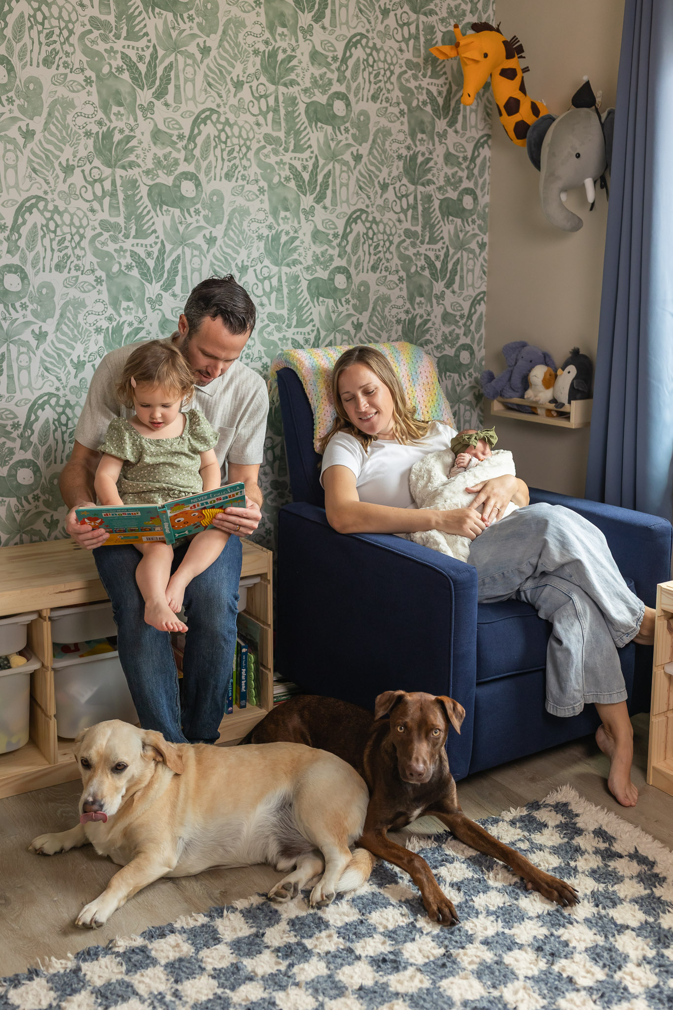 A man with two children and two dogs in a living room with patterned wallpaper and animal wall decorations.