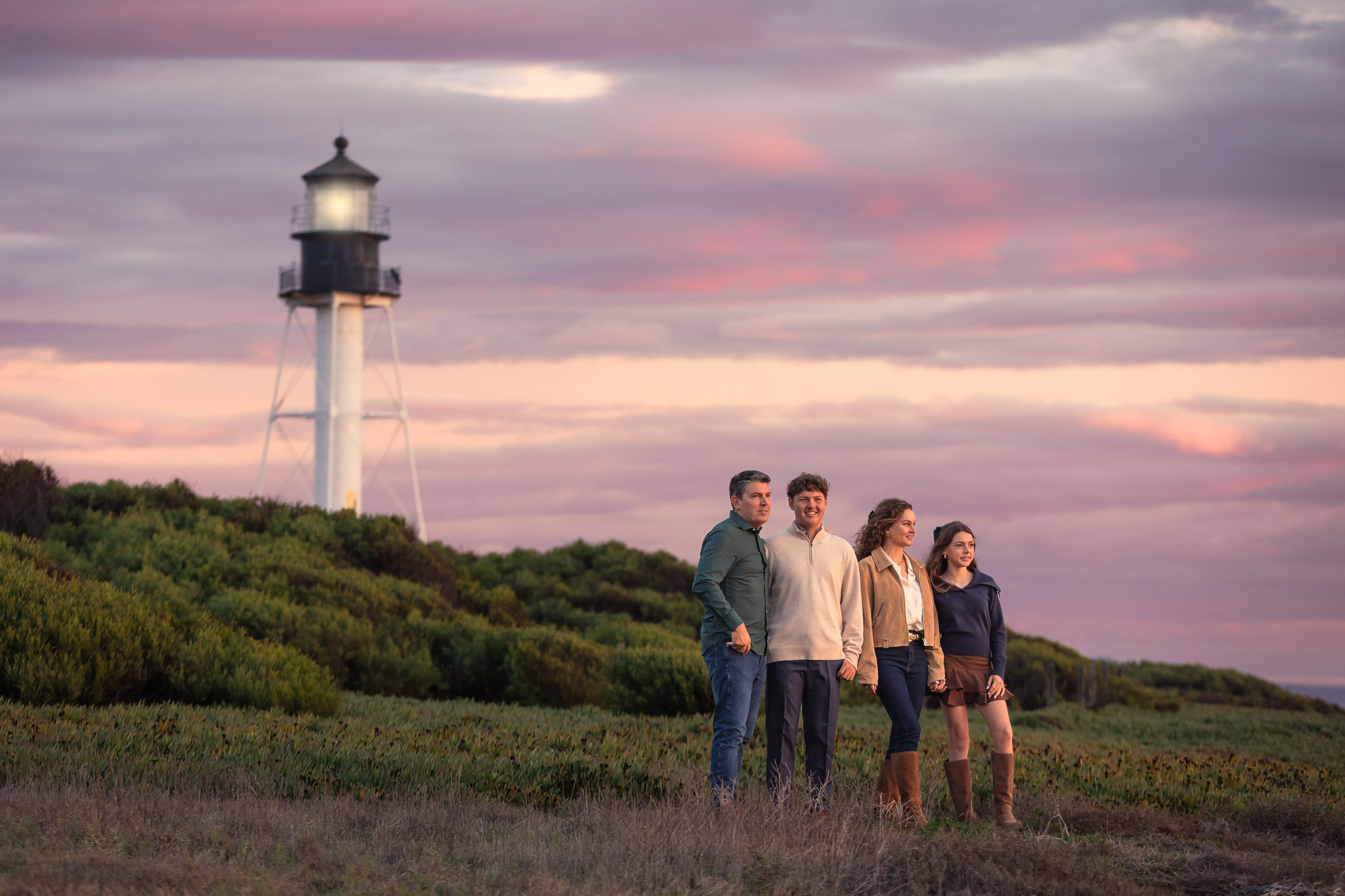 Four people walking along a grassy area near a lighthouse with a colorful sky background.