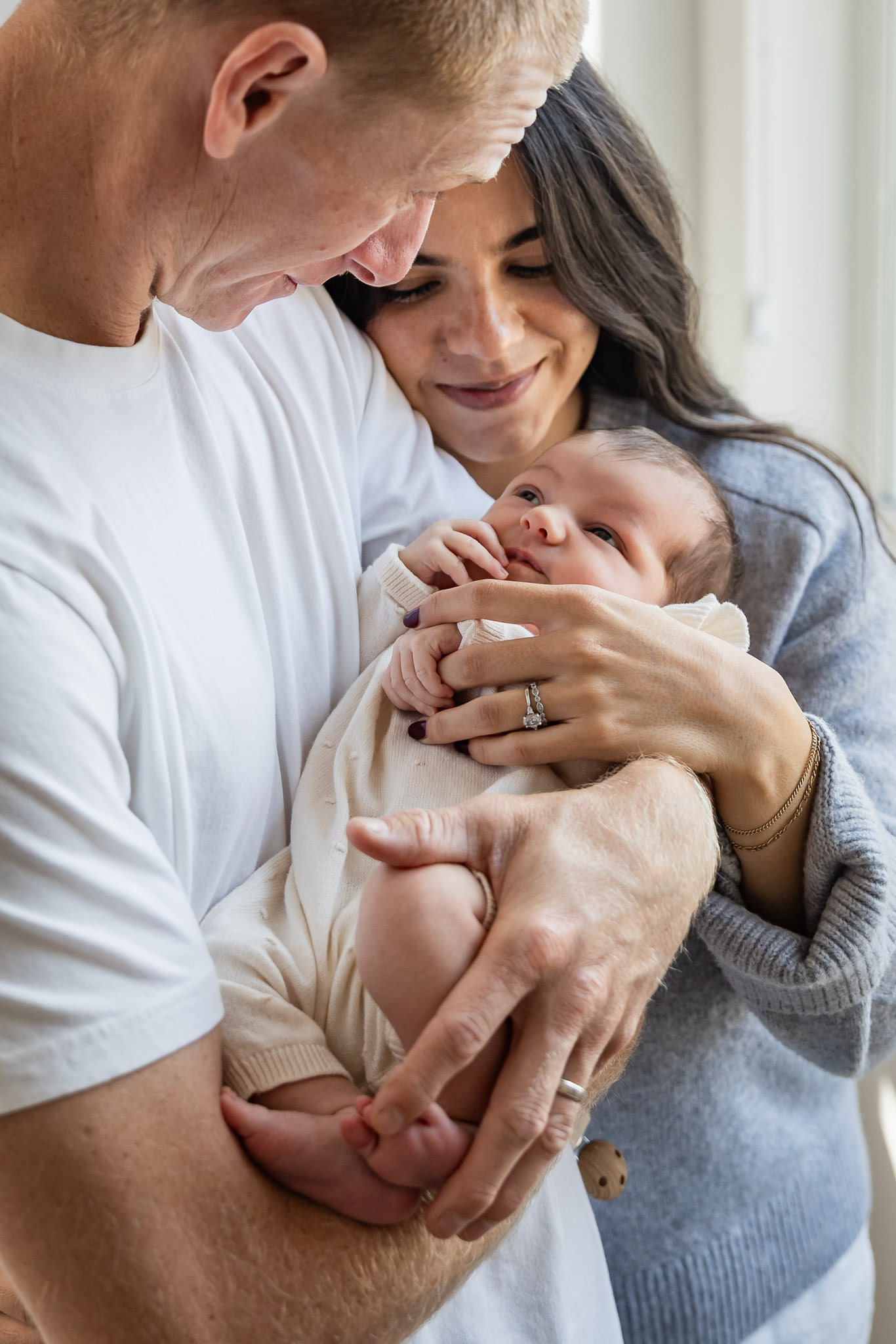 Family holding a baby, smiling and looking at each other, indoors near a window.