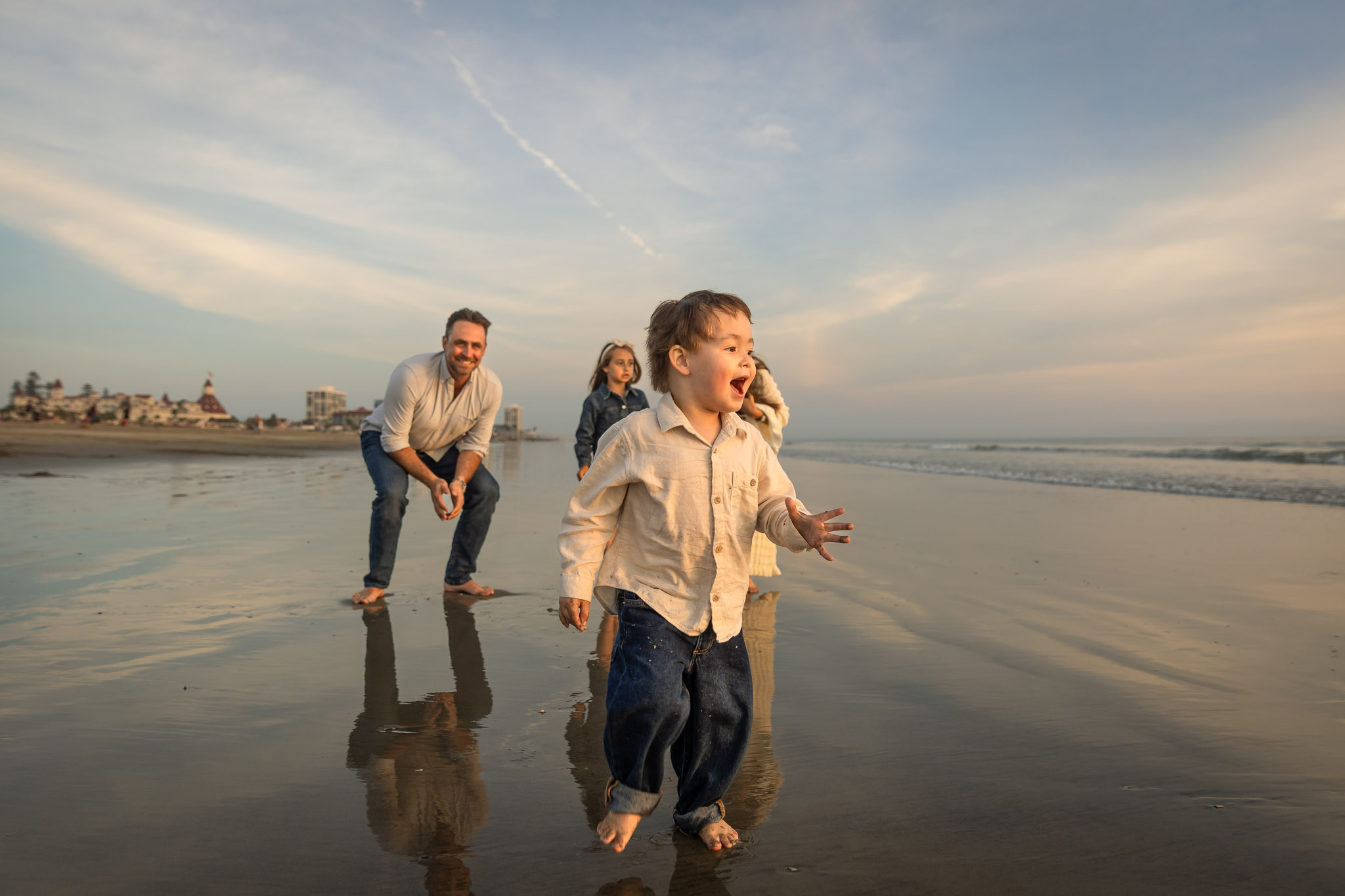 Child running on the beach with three people in the background, ocean and sky visible, reflections on wet sand, daytime.