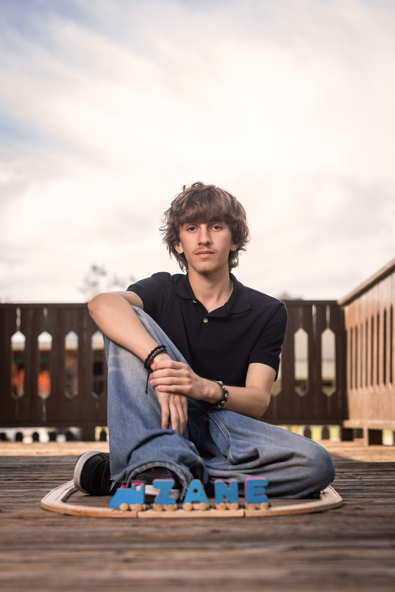 Young man with curly hair sitting on a wooden deck, with a fence and sky in the background.
