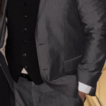 Man in a gray suit and white shirt smiling, standing indoors with plants in the background.