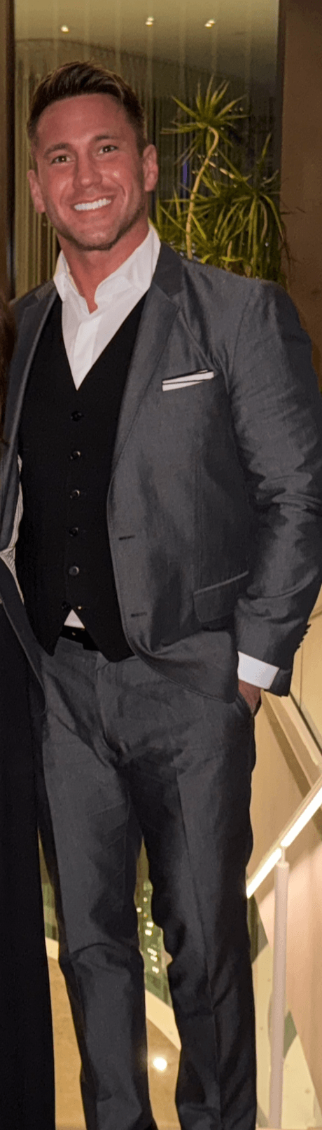Man in a gray suit and white shirt smiling, standing indoors with plants in the background.