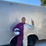 Person standing next to a trailer, holding a helmet, waving, with a clear sky in the background.