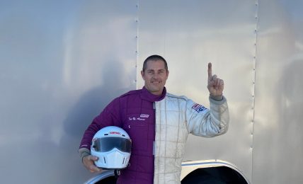 Person standing next to a trailer, holding a helmet, waving, with a clear sky in the background.