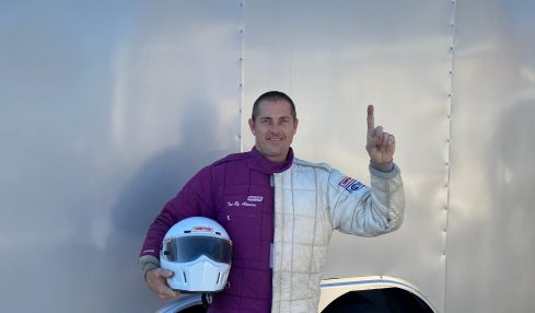 Person standing next to a trailer, holding a helmet, waving, with a clear sky in the background.