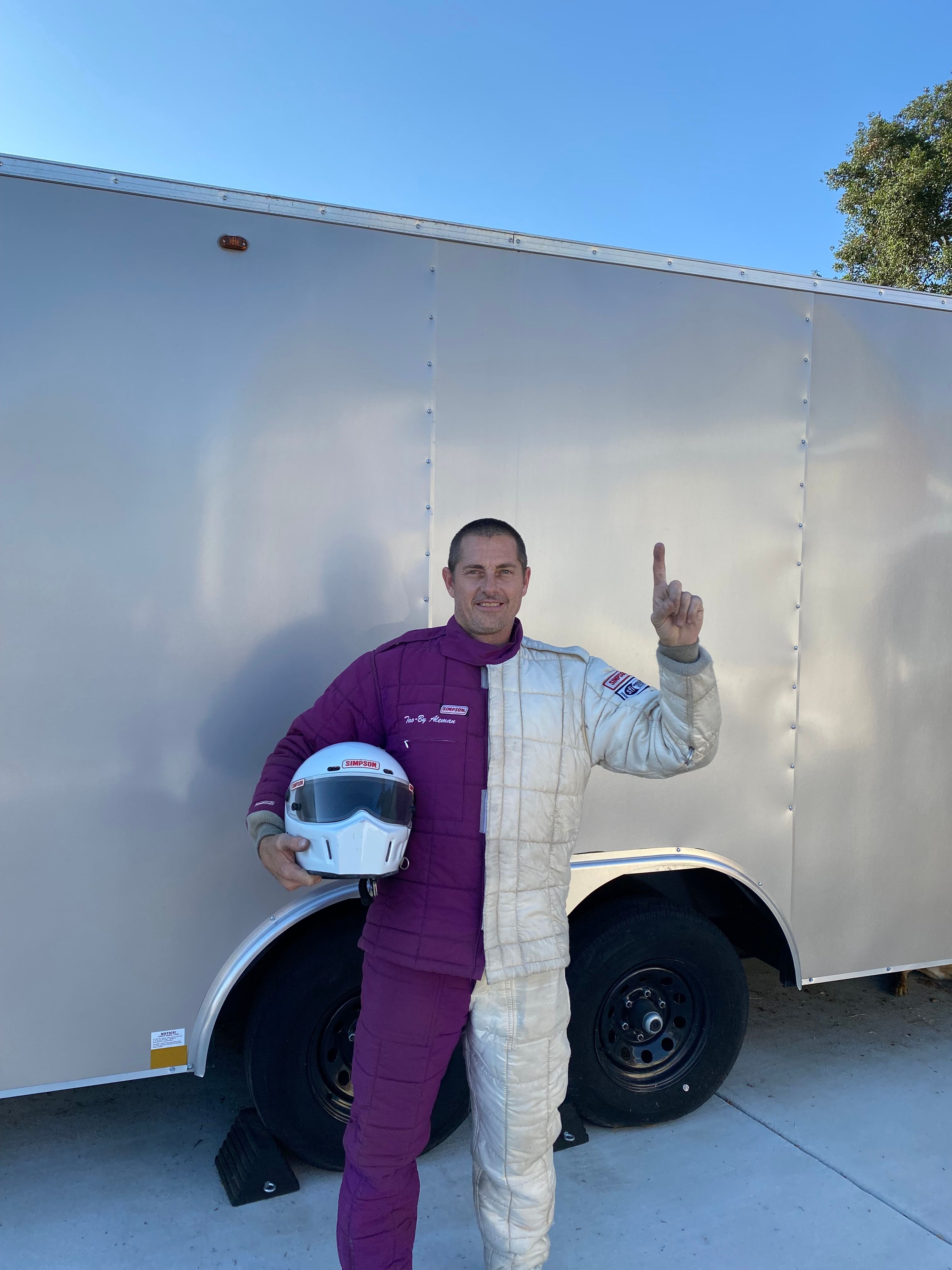 Person standing next to a trailer, holding a helmet, waving, with a clear sky in the background.