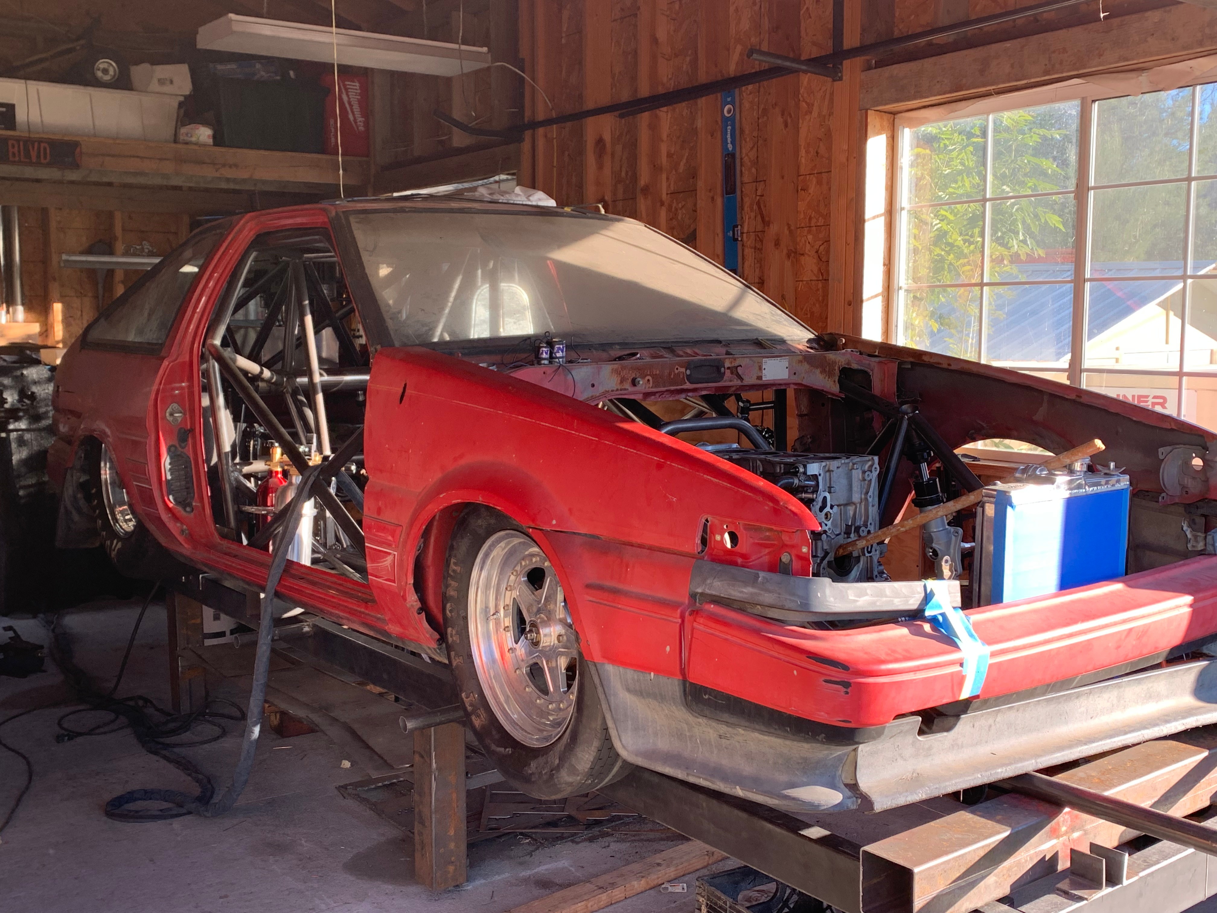 Red race car inside a wooden garage, with open hood and visible engine components, on a lift with tools nearby.
