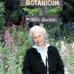 Older woman with white hair standing in a garden with pink and purple flowers, smiling, with signs for Theatrum Botanicum and Hills Garden behind her.