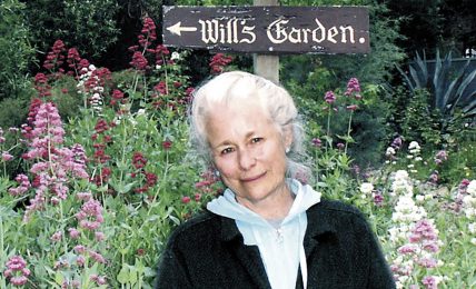Older woman with white hair standing in a garden with pink and purple flowers, smiling, with signs for Theatrum Botanicum and Hills Garden behind her.
