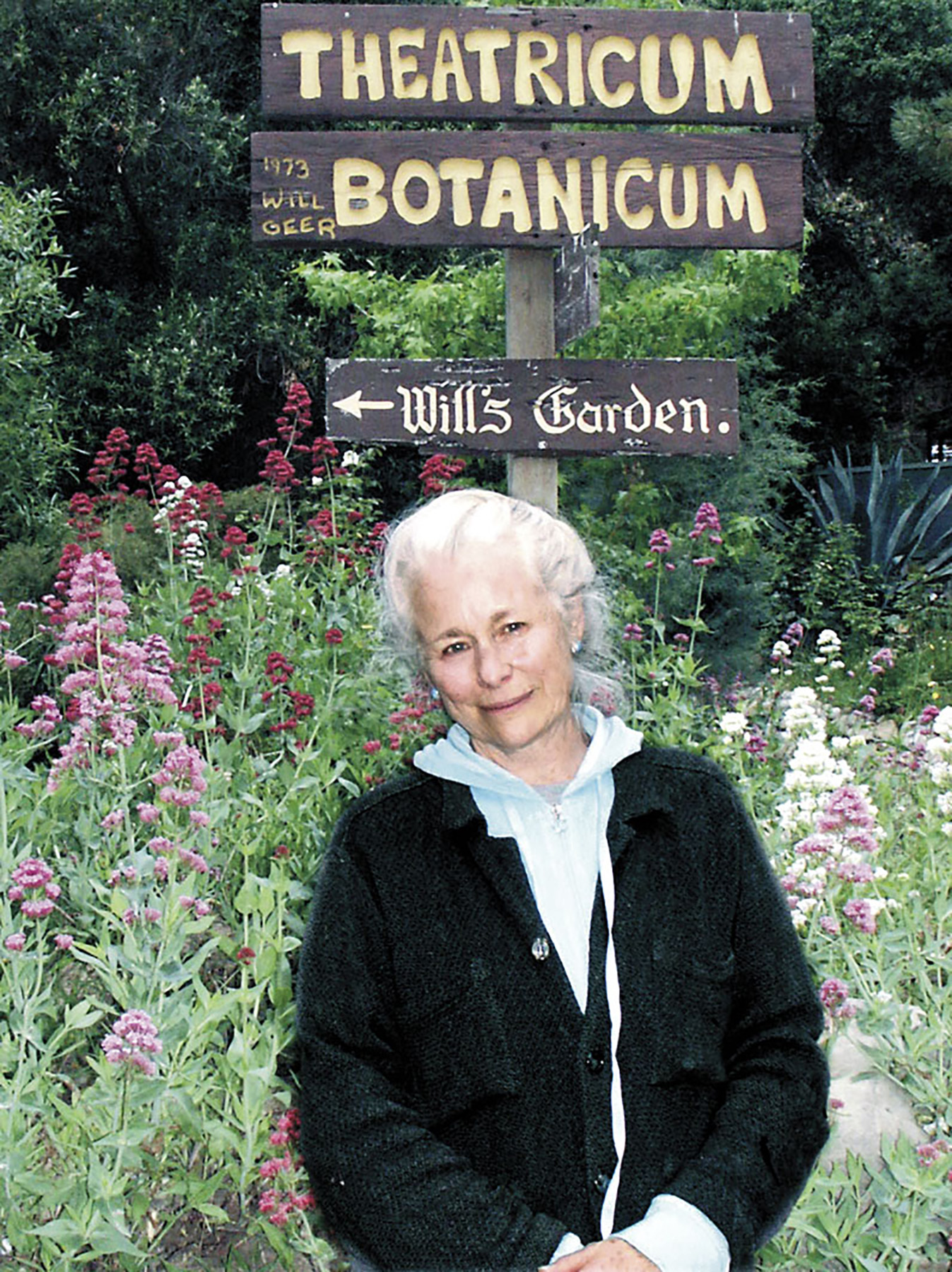 Older woman with white hair standing in a garden with pink and purple flowers, smiling, with signs for Theatrum Botanicum and Hills Garden behind her.