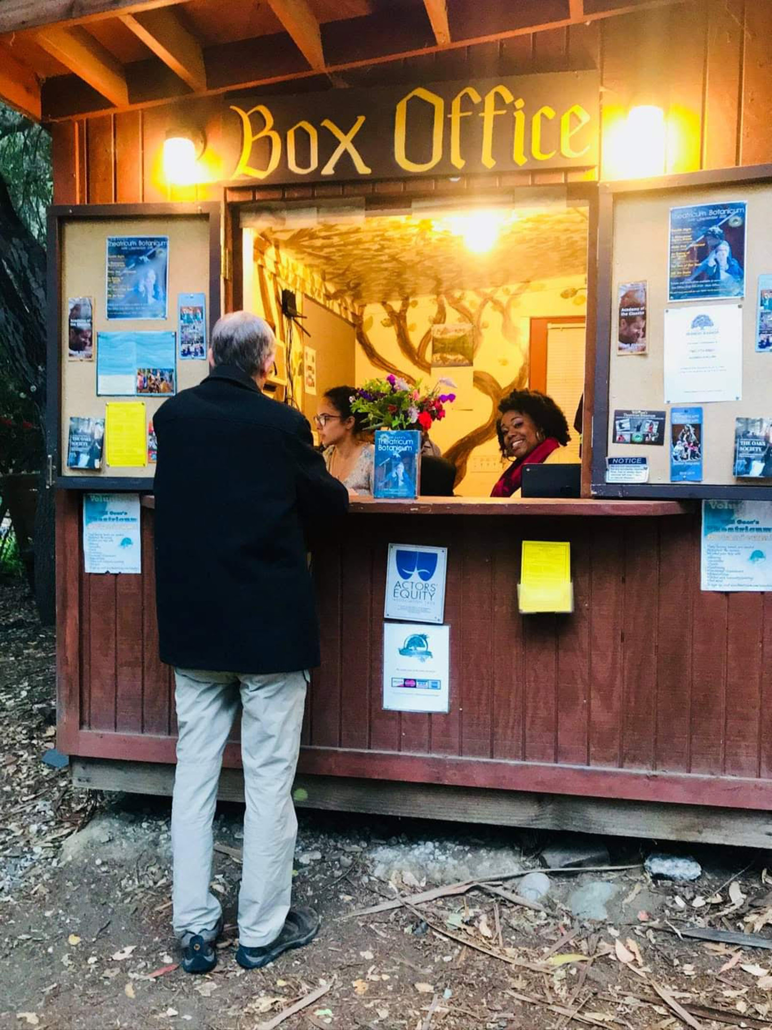 Person at box office counter talking to staff, posters on sides, warm lighting, outdoor setting, flowers on counter.