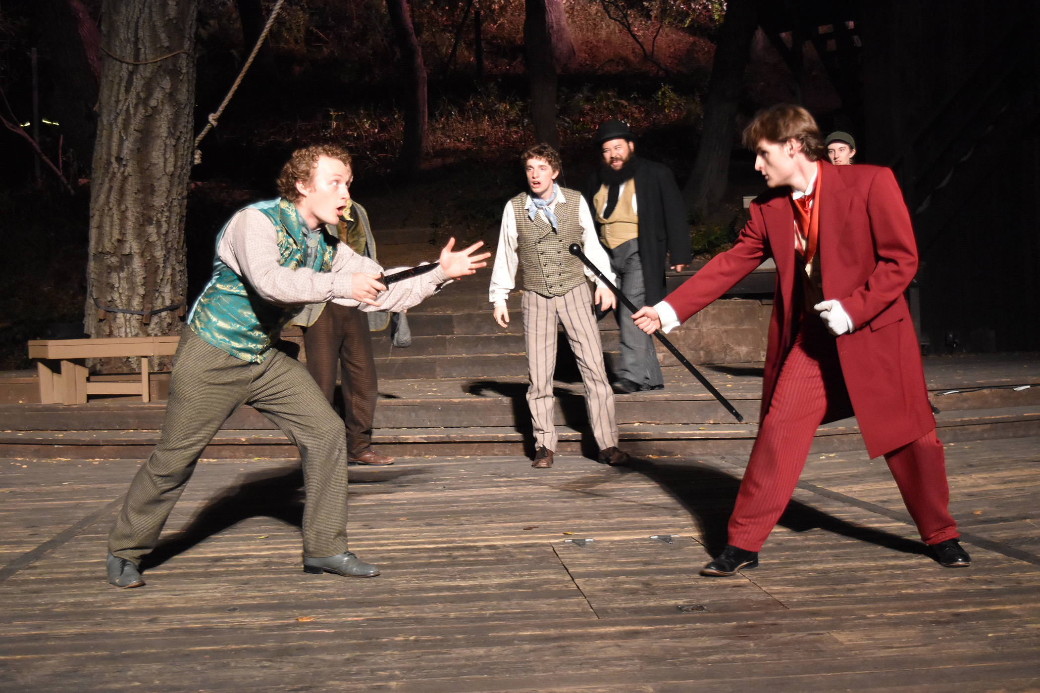 Two actors face off with swords on stage, surrounded by three other actors watching, outdoors with trees in background.