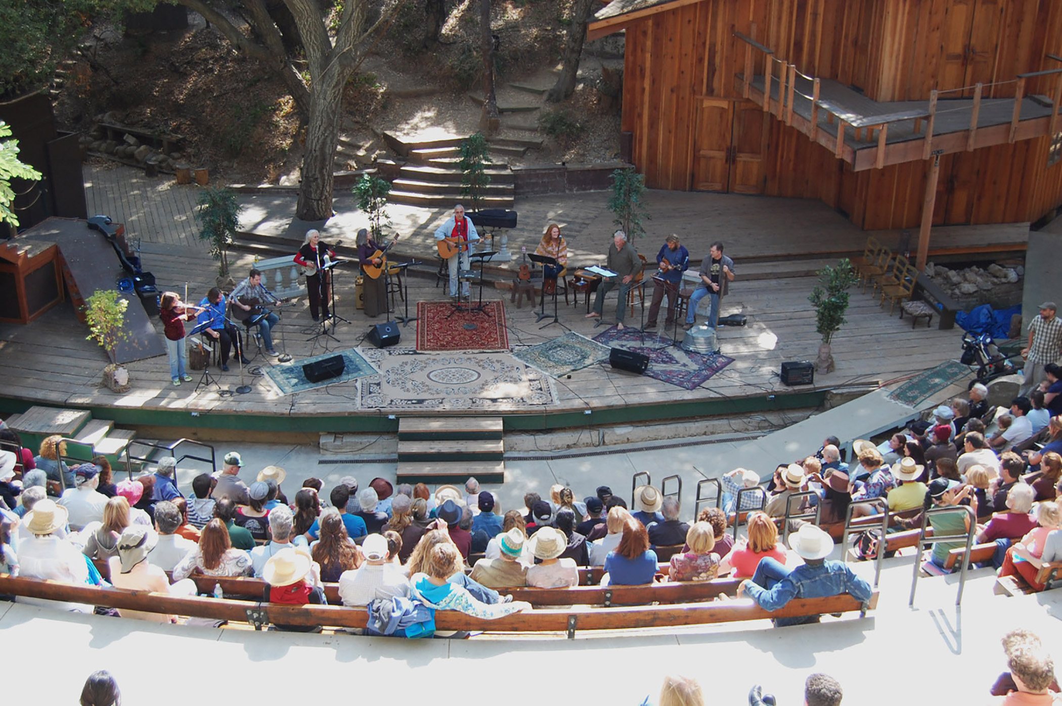 Outdoor stage with musicians performing, audience seated in semi-circle, trees and wooden building in background.
