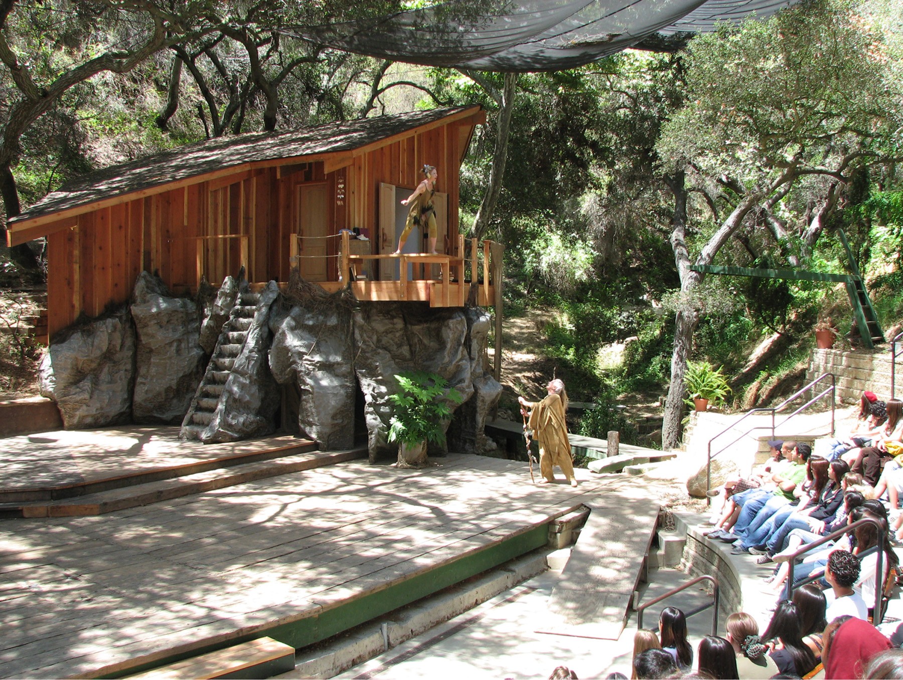 Outdoor theater with a wooden stage, audience seated, and trees in the background.