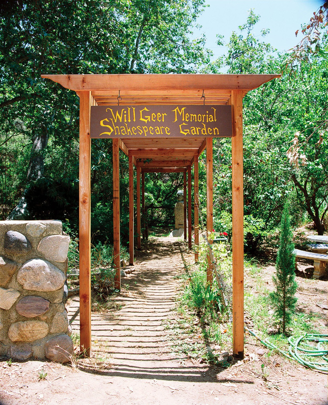 Wooden walkway with a sign reading 'Wall Geer Memorial Shakespeare Garden' surrounded by trees and plants.