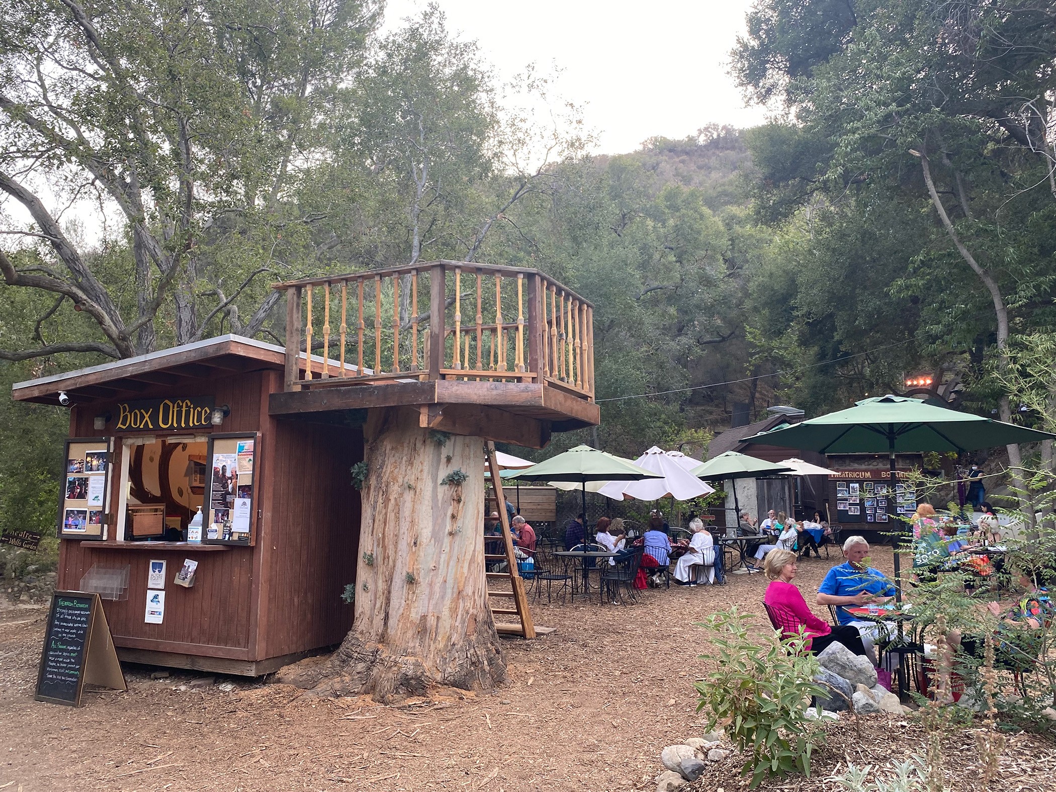 Outdoor scene with a small wooden booth, trees, and people sitting at tables with umbrellas.