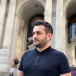 Man with short dark hair and beard wearing a black polo shirt standing outside a historic building with arches.