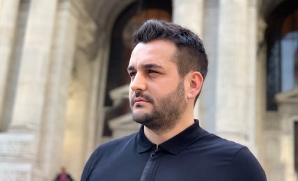 Man with short dark hair and beard wearing a black polo shirt standing outside a historic building with arches.