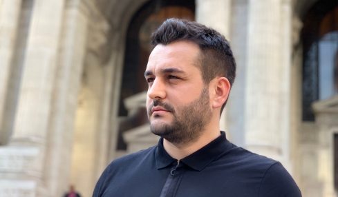 Man with short dark hair and beard wearing a black polo shirt standing outside a historic building with arches.