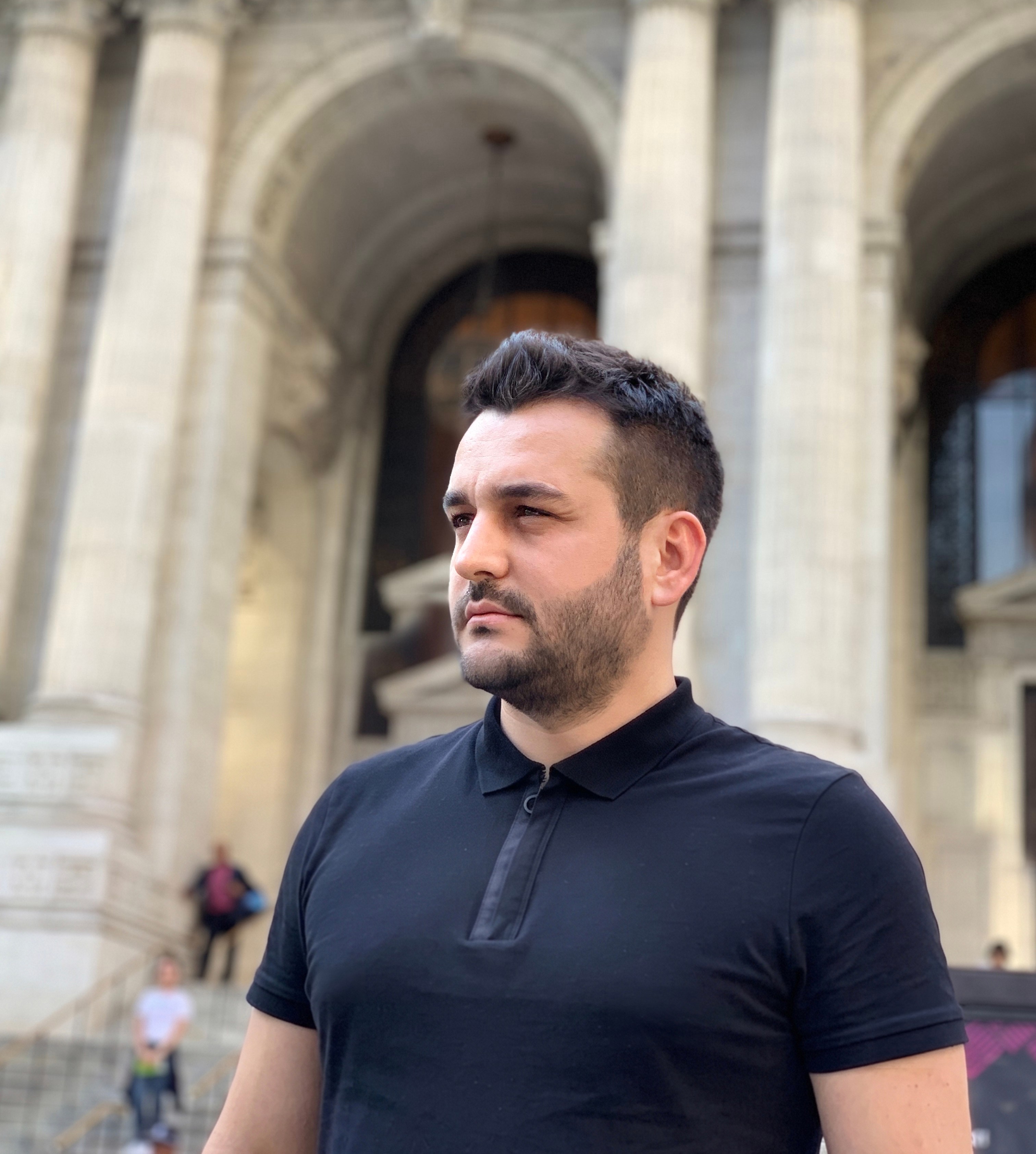 Man with short dark hair and beard wearing a black polo shirt standing outside a historic building with arches.