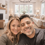Two people smiling in a bright living room with large windows, a chandelier, and cozy furniture.