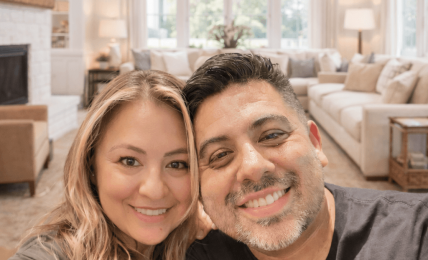 Two people smiling in a bright living room with large windows, a chandelier, and cozy furniture.