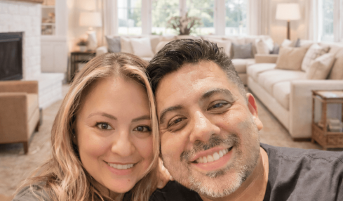 Two people smiling in a bright living room with large windows, a chandelier, and cozy furniture.