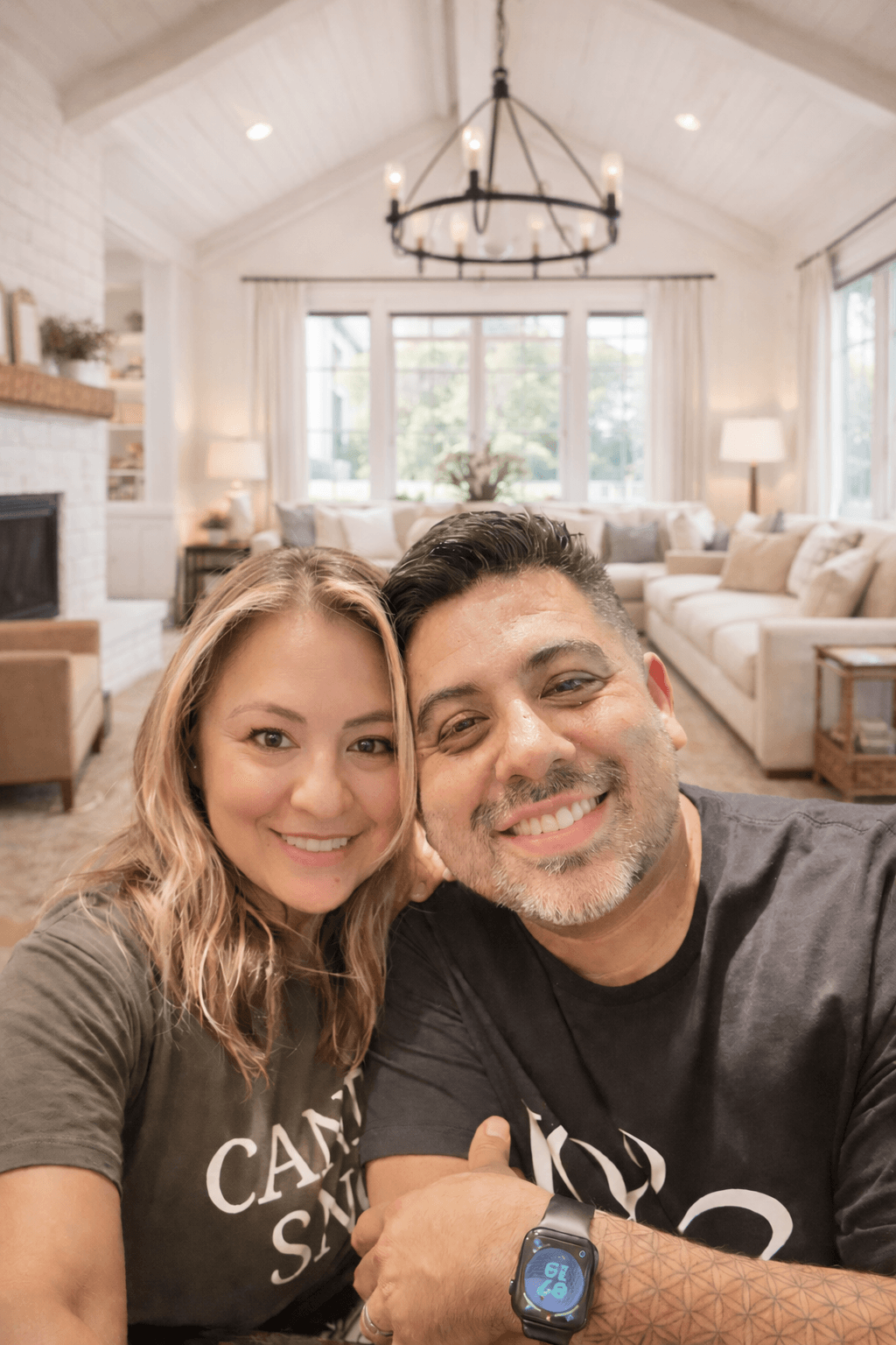 Two people smiling in a bright living room with large windows, a chandelier, and cozy furniture.