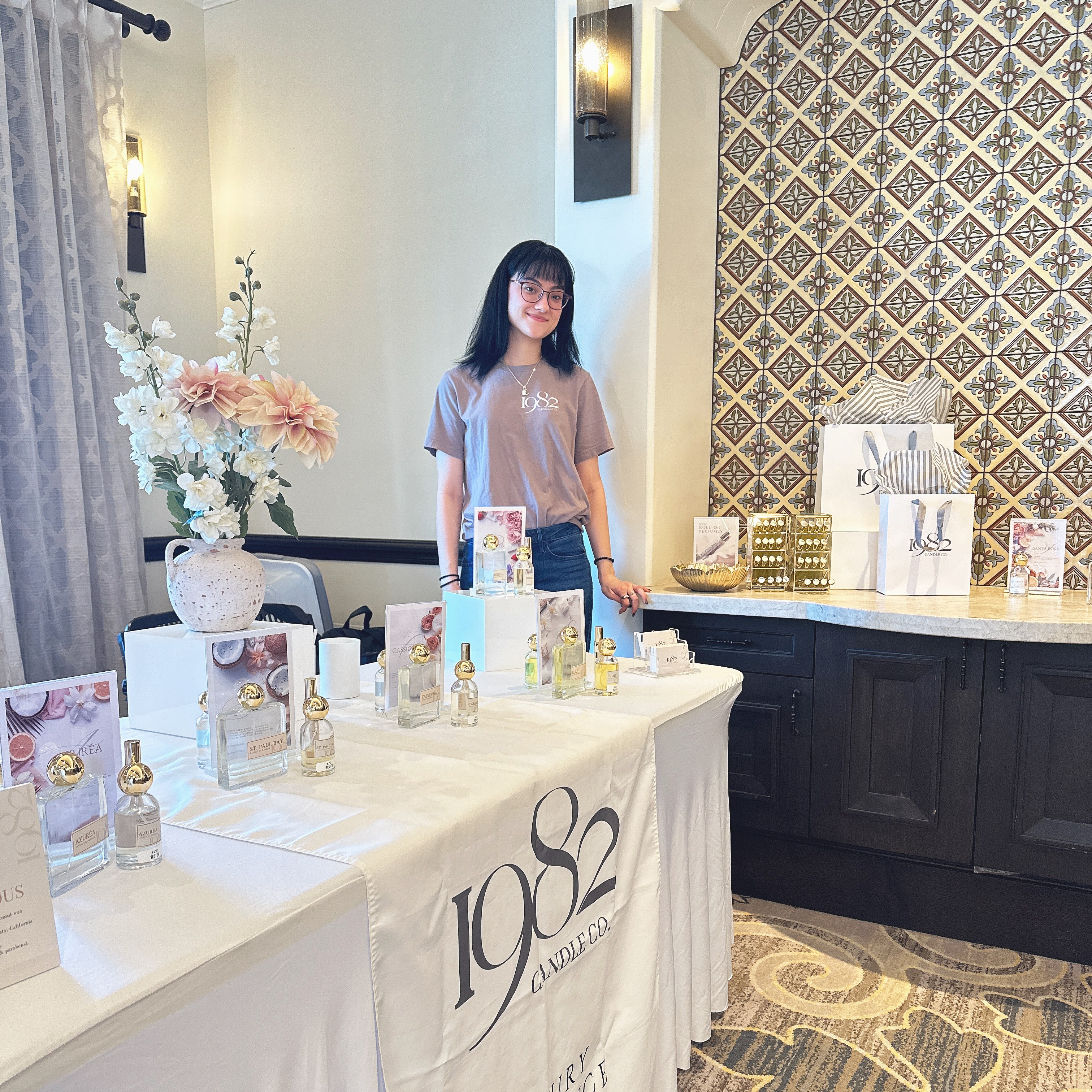 Young woman with glasses standing behind a table with gift items in a decorated room.