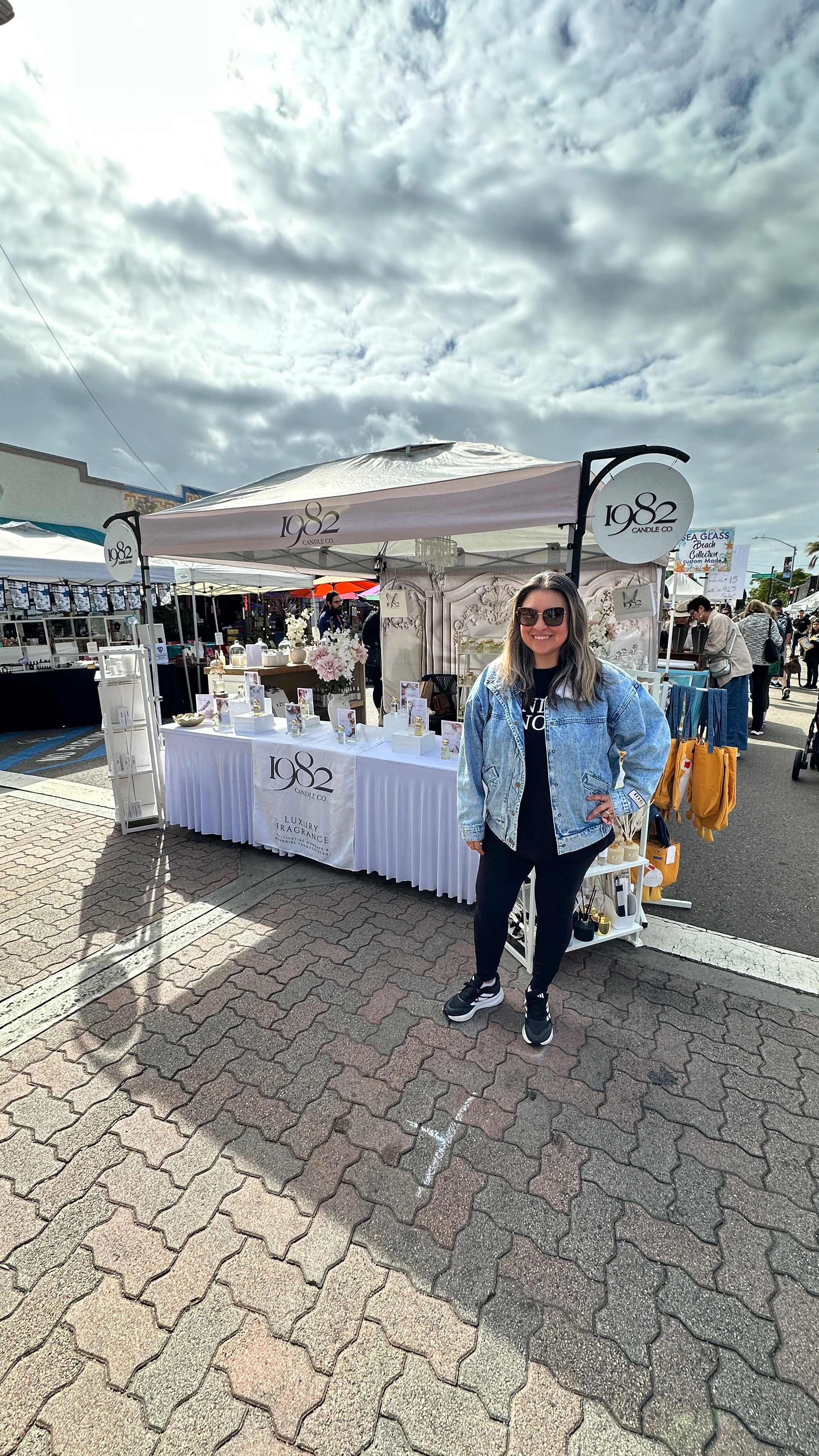 Woman standing in front of a booth at an outdoor market, with cloudy sky overhead.