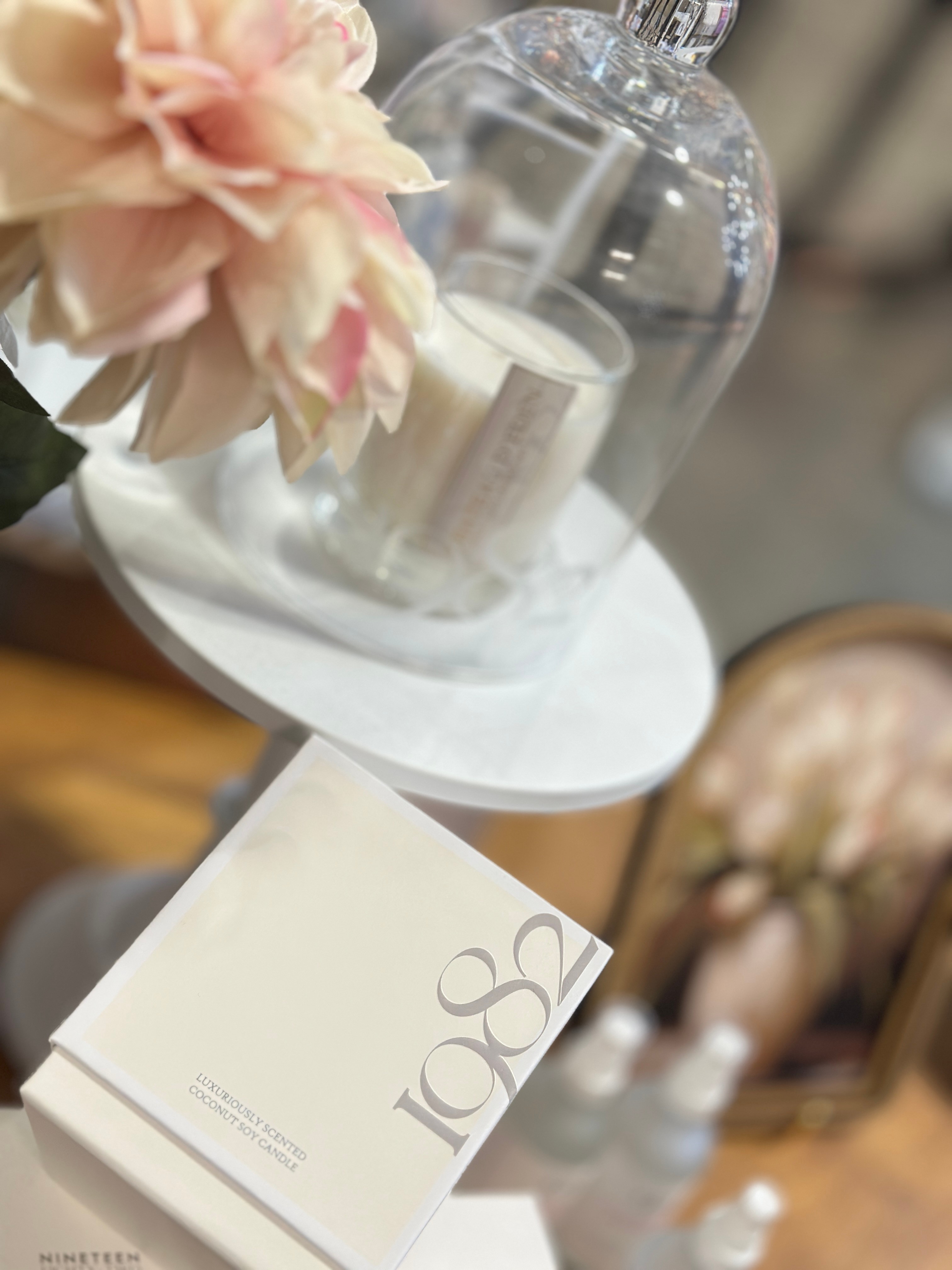 A glass cloche covers a candle and flower arrangement on a white table, with a white box nearby. Blurred background.