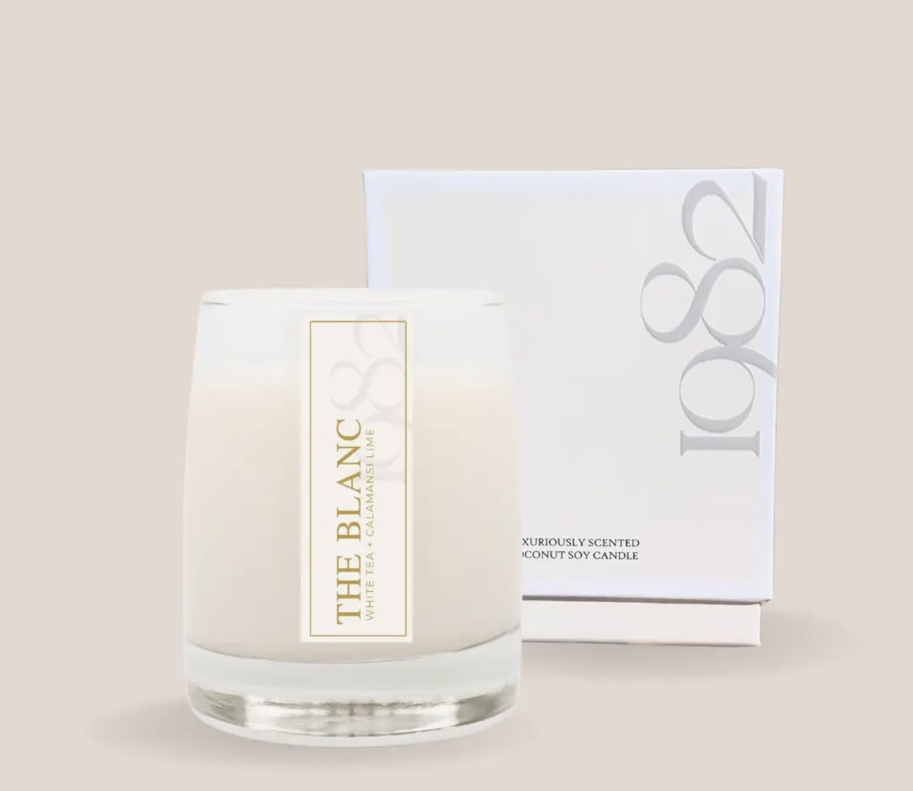 A white scented candle in a glass container with a label, placed in front of a white box with gray text.