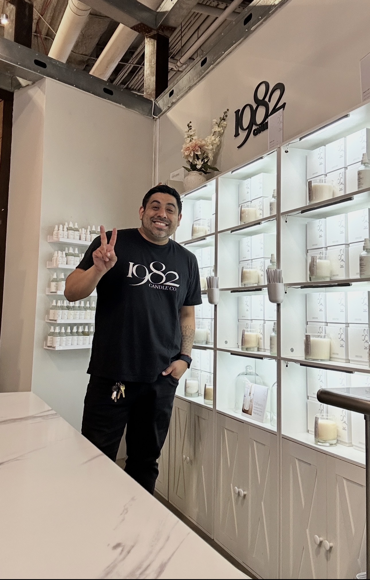 Smiling man making a peace sign in a store with shelves of candles and decor, wearing a black T-shirt with white text.