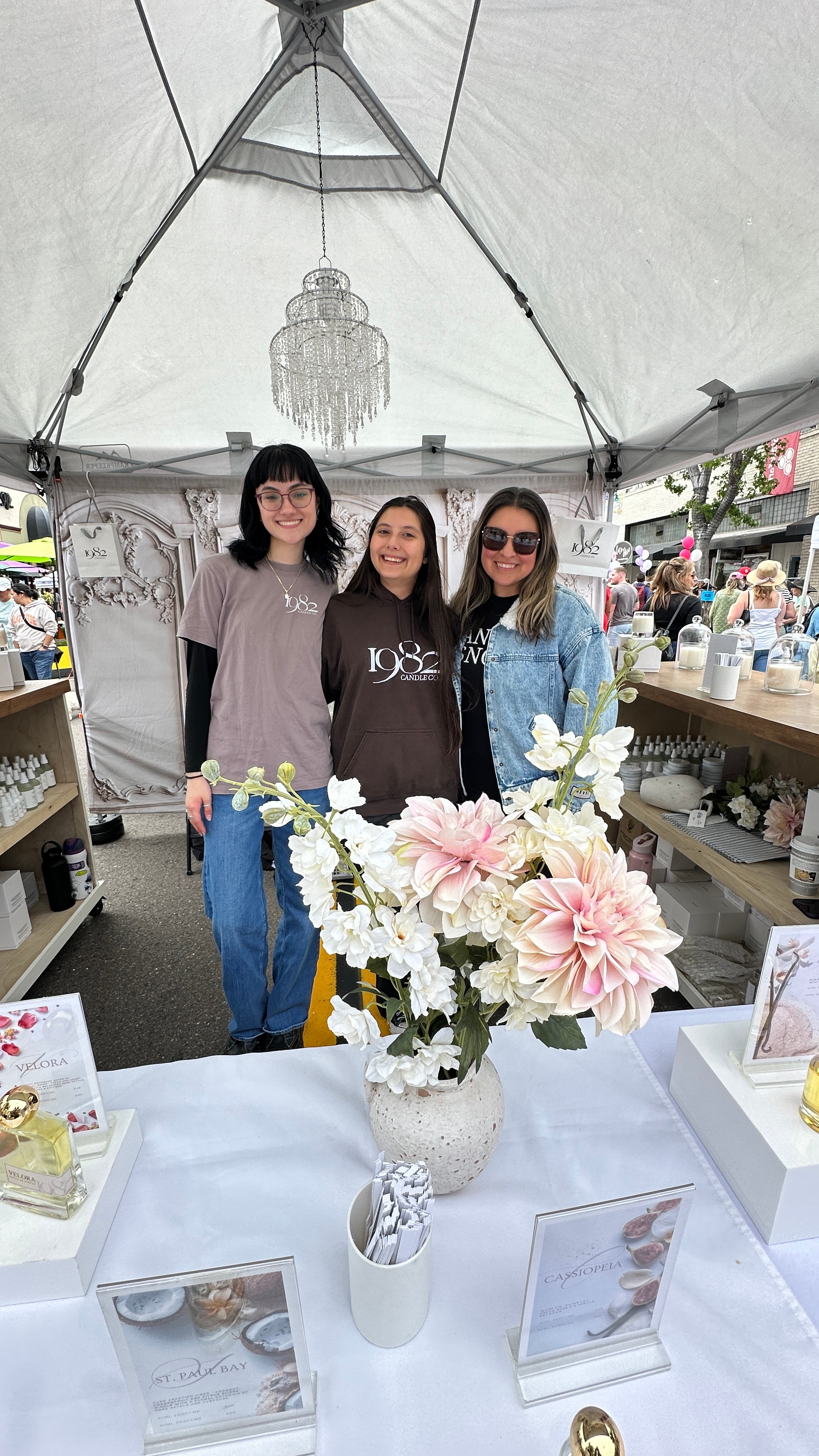 Four women stand inside a tent behind a table with a large flower arrangement, smiling at the camera.