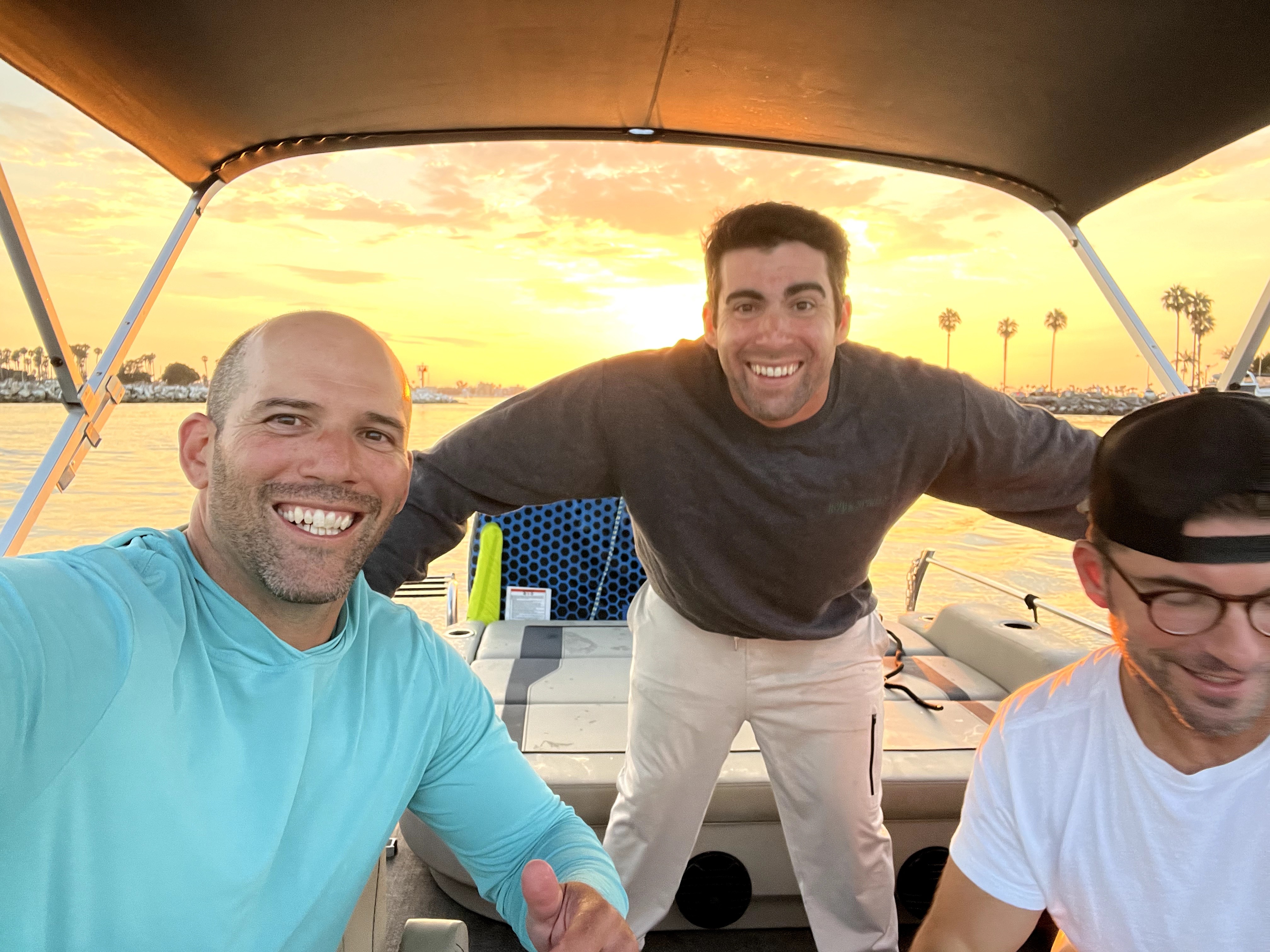 Three men smiling on a boat during sunset, with water and palm trees in the background.