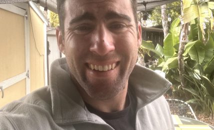 Smiling man outdoors with plants and a canopy overhead, wearing a light gray jacket and black shirt.