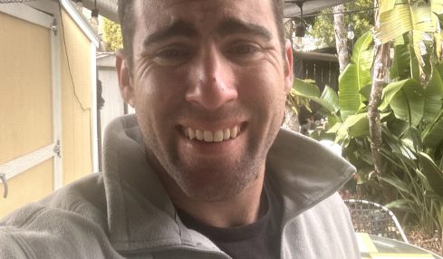 Smiling man outdoors with plants and a canopy overhead, wearing a light gray jacket and black shirt.