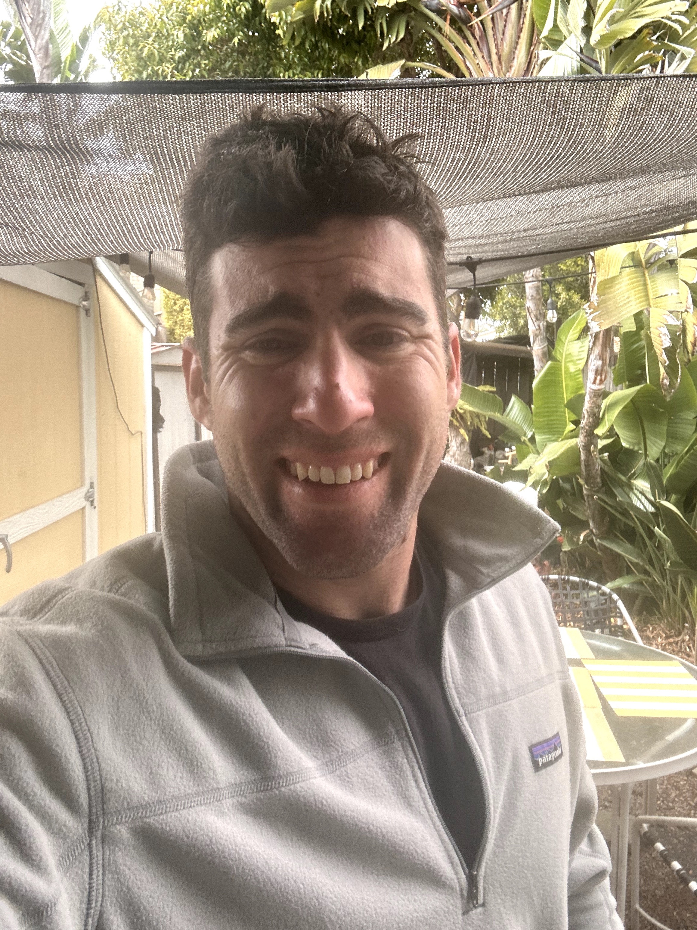 Smiling man outdoors with plants and a canopy overhead, wearing a light gray jacket and black shirt.